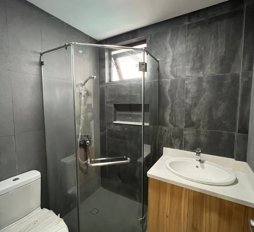 Ensuite bathroom finished in dark charcoal tile, with a frameless glass shower enclosure, handheld shower head, small window, and a wood vanity with an oval white sink