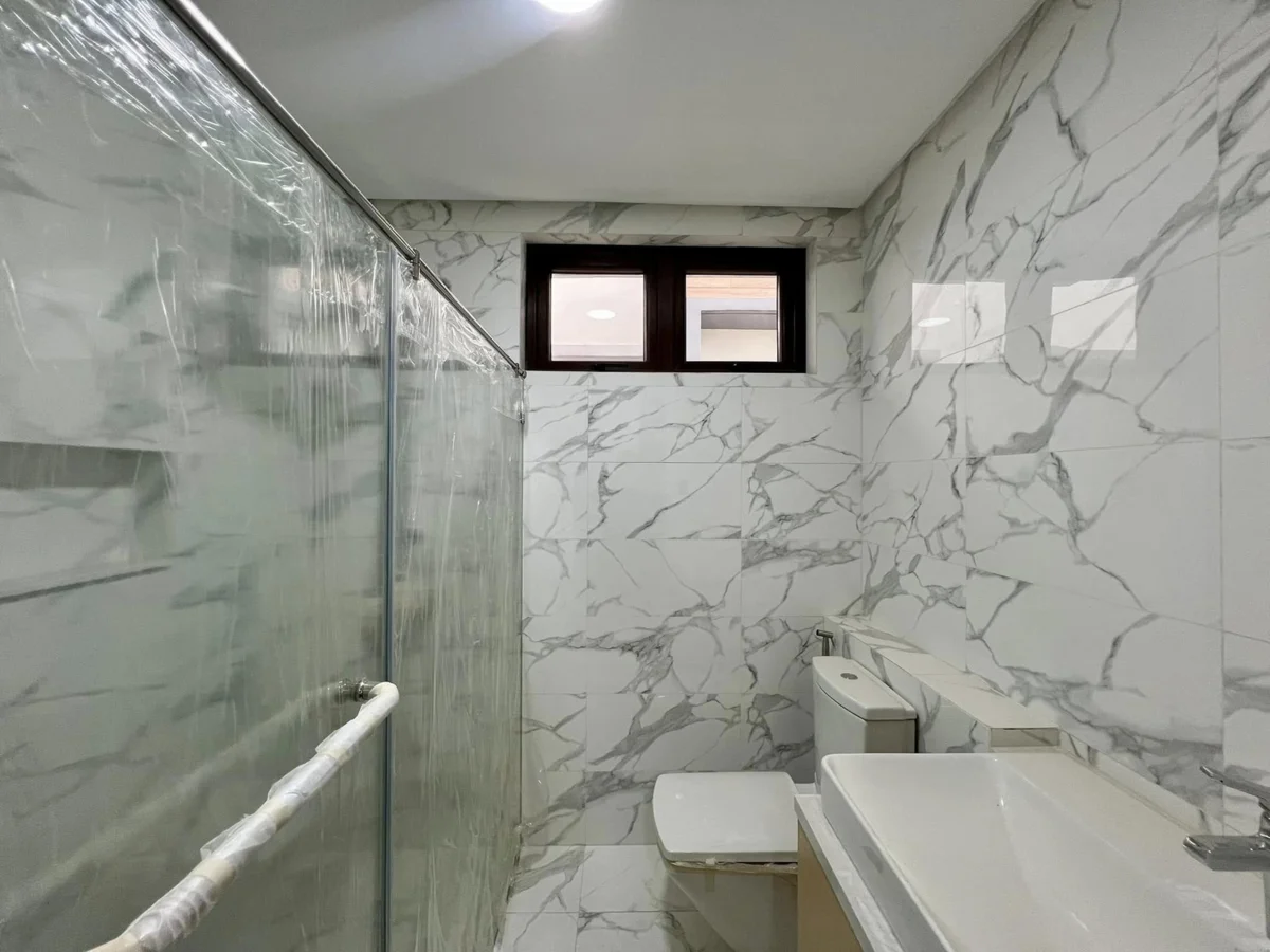 Bathroom finished in white-and-gray marble-look tile, with a plastic-wrapped glass shower panel, dark wood-framed window, toilet, and a white vessel sink on a wood-tone counter
