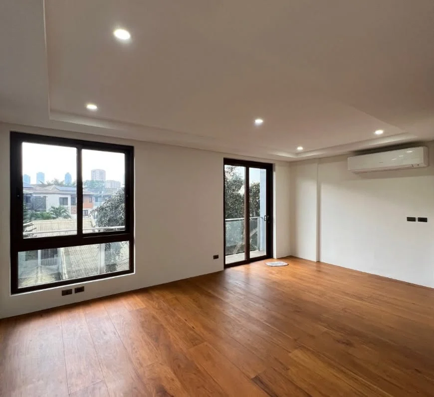 Empty bedroom with wood plank flooring, a wall-mounted air conditioner, and a sliding glass door opening onto a small balcony with a view of the Quezon City skyline