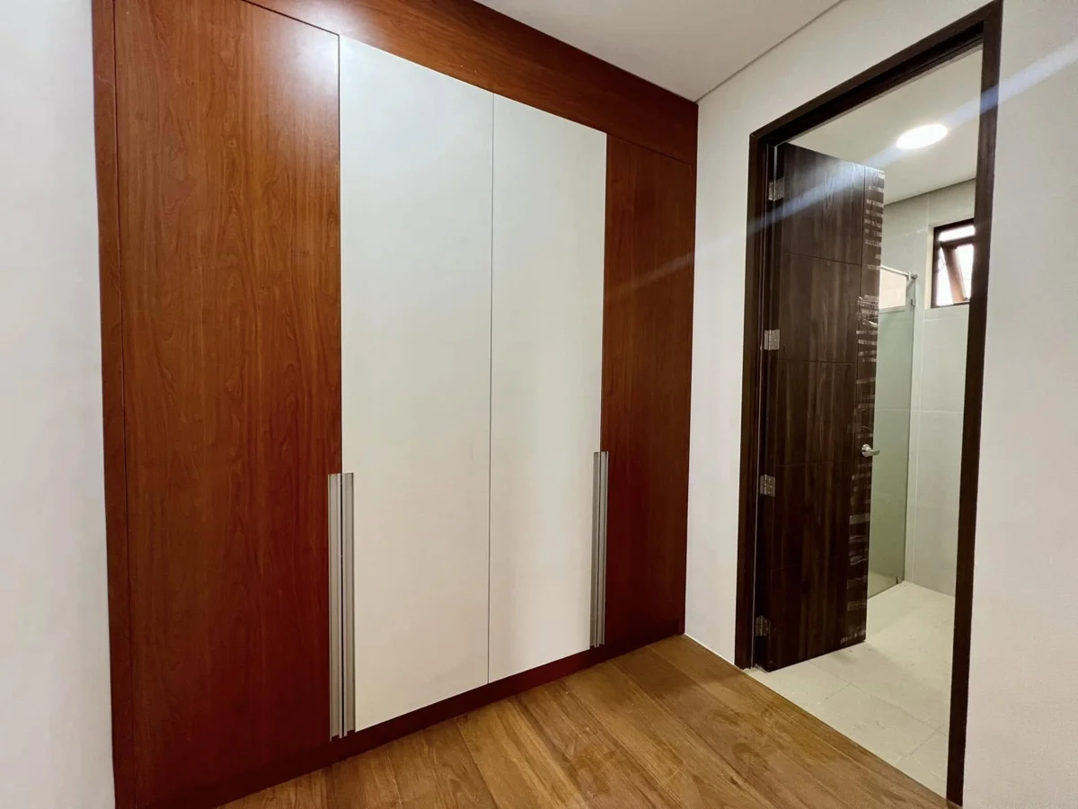 Bedroom built-in wardrobe with wood-grain and white paneled doors and brushed-metal handles, next to an open dark-wood door leading into an ensuite bathroom with a glass shower