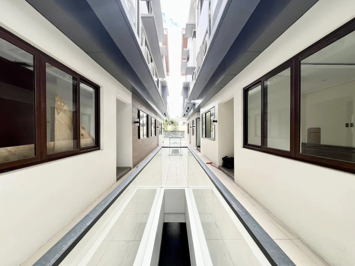 Second-level courtyard corridor with a glass floor panel let into the walkway, doors and dark-wood-framed windows lining both sides, open sky visible overhead