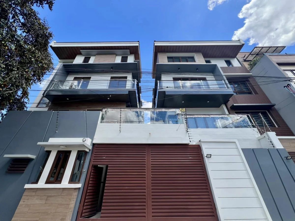 Street-level view looking up at the twin townhouse facade, with dark gray upper stories, wood-look eaves, glass balcony railings, and a maroon slatted garage door at ground level