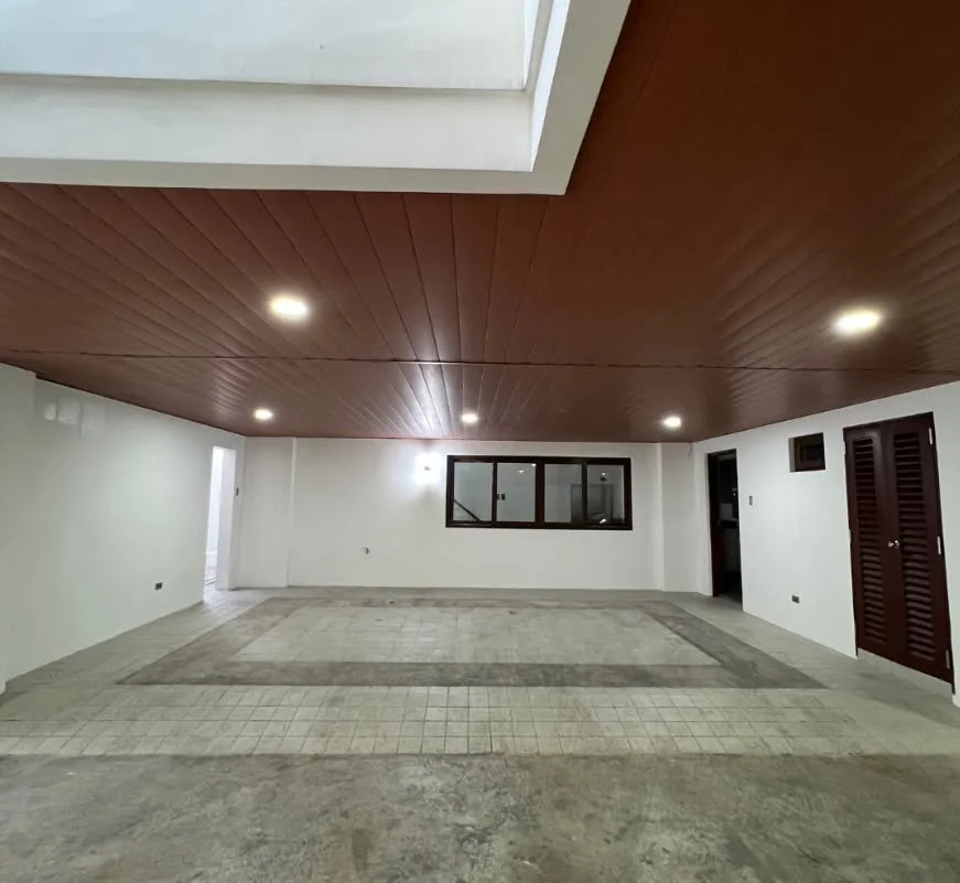Covered ground-floor garage bay with a dark reddish-brown paneled ceiling, bare concrete-and-tile floor, a wood louvered double door, and a window into an adjoining room