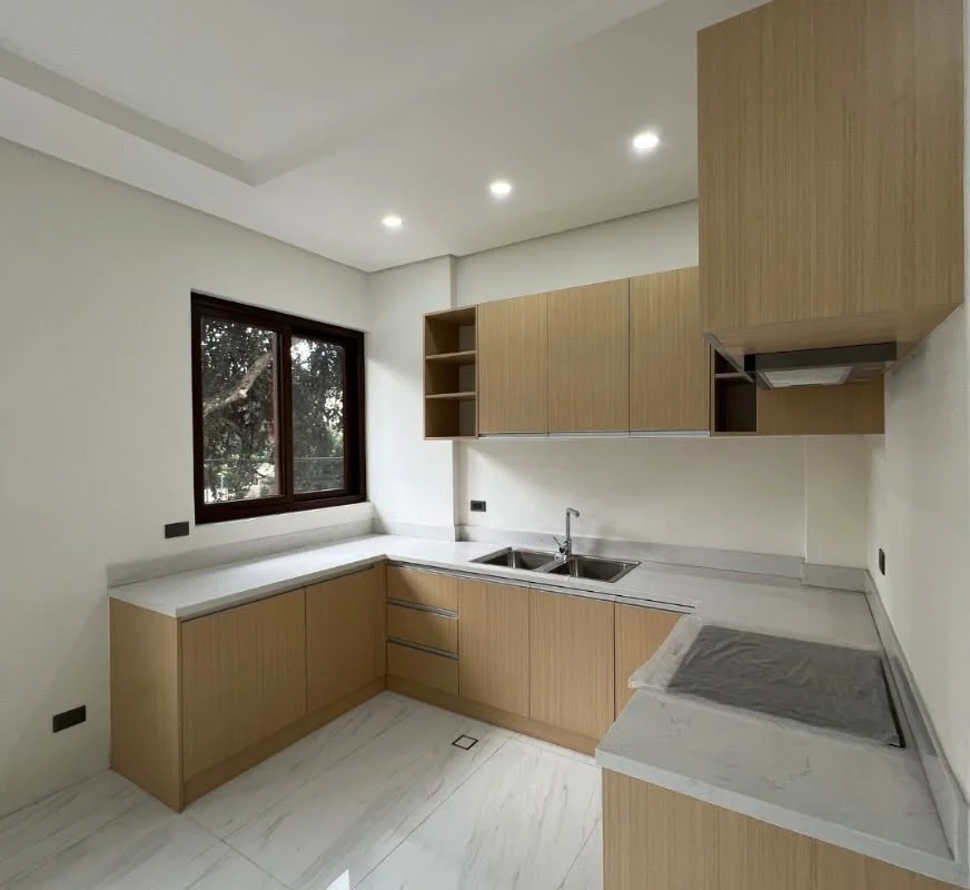 Kitchen with light wood cabinetry, gray marble-look countertops, a double-basin sink below a window, and a stovetop area still covered in plastic sheeting