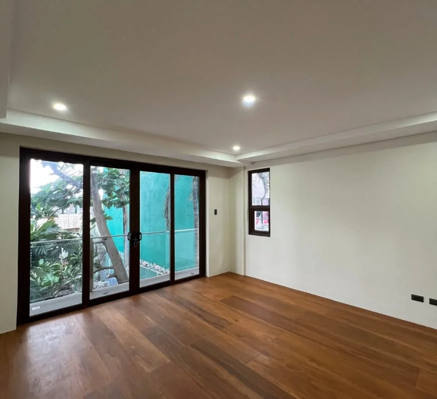 Empty living space with wood plank flooring and a wood-framed sliding glass door opening onto a balcony with a glass-and-metal railing, greenery and a neighboring teal-painted wall visible outside
